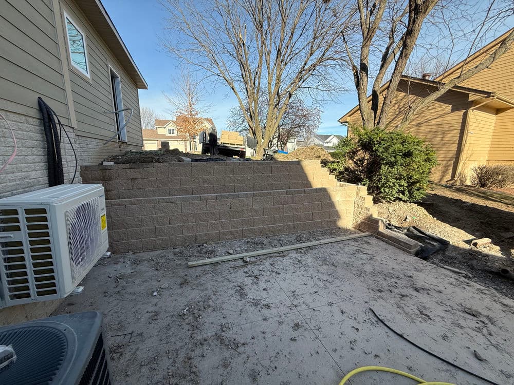 Image of a construction site showing a stone retaining wall, air conditioning unit, and nearby trees.