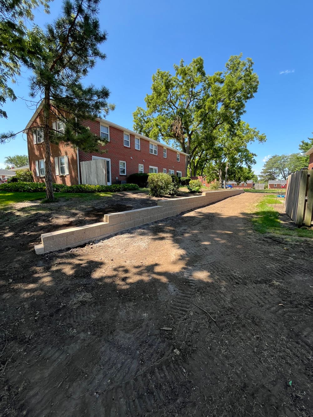 Newly paved pathway next to brick apartment building surrounded by trees and grass.