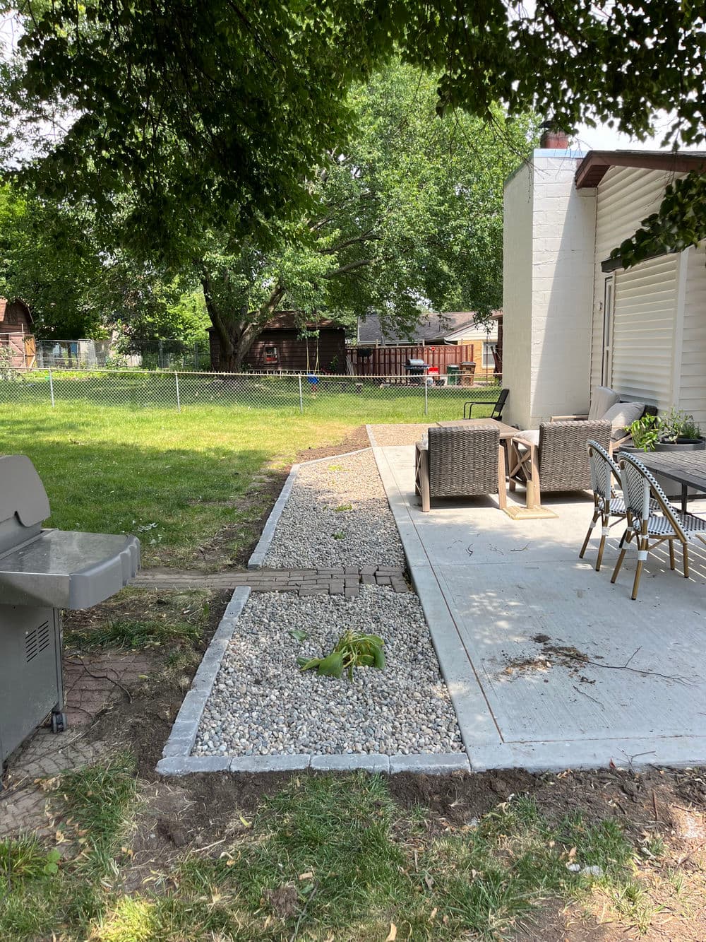 Backyard patio with gravel pathway, seating area, grill, and lush green trees.