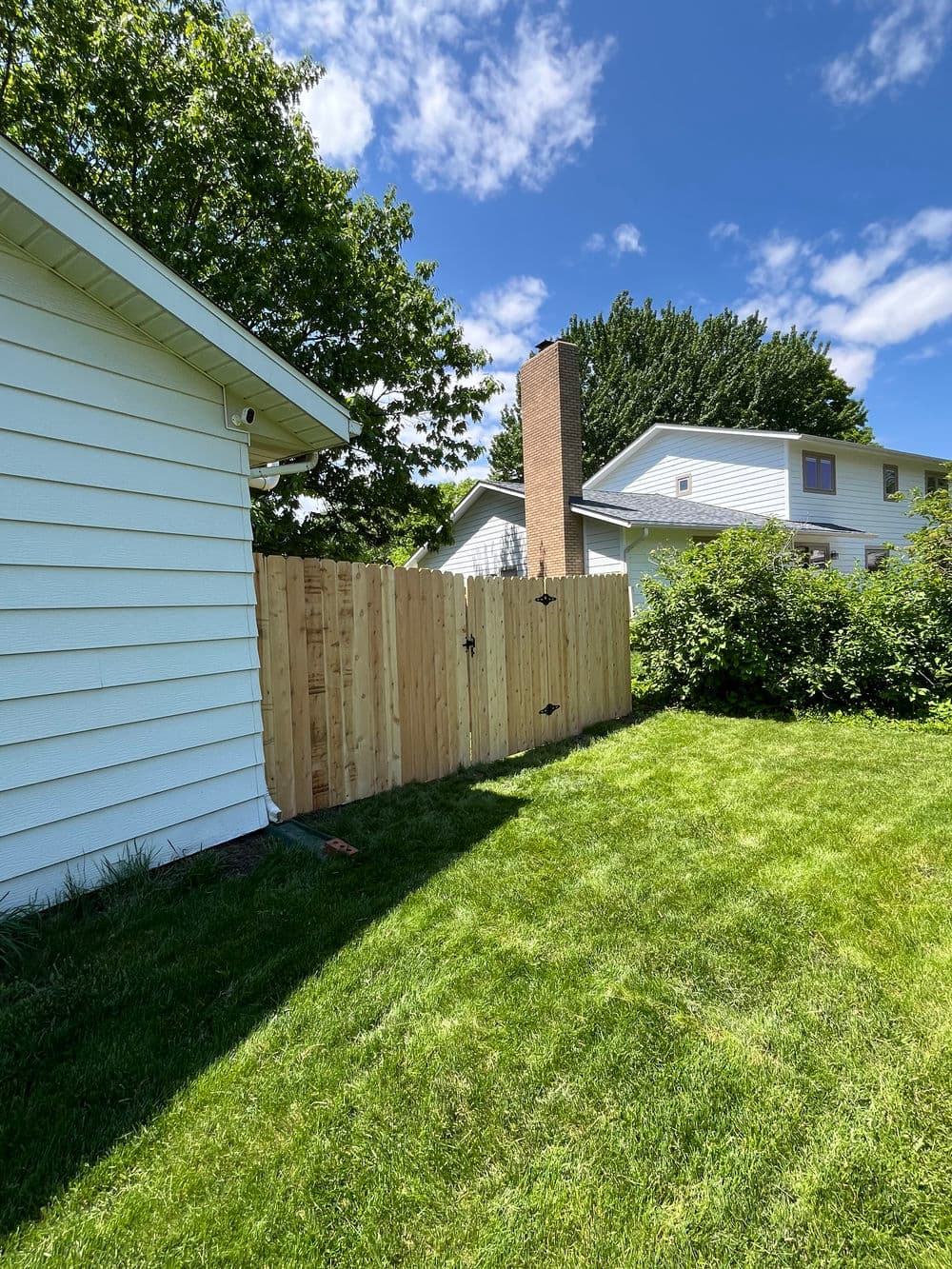 Wooden fence partially shaded by trees in a green backyard with two houses in the background.
