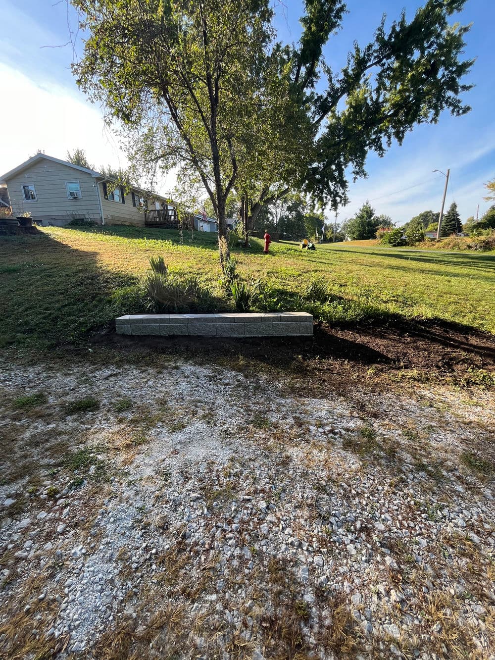 Landscaped yard with a retaining wall, tree, and green grass under a clear blue sky.