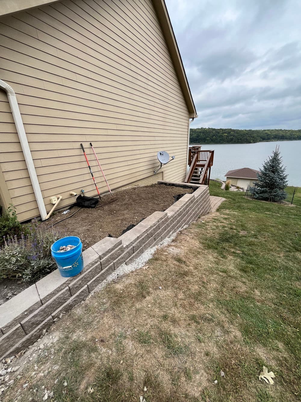 Landscape preparation near lakefront home with gravel and retaining wall, cloudy sky above.