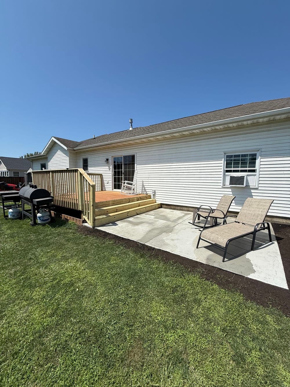 Backyard patio with deck, grill, and lounge chairs under clear blue sky. Ideal for relaxation.