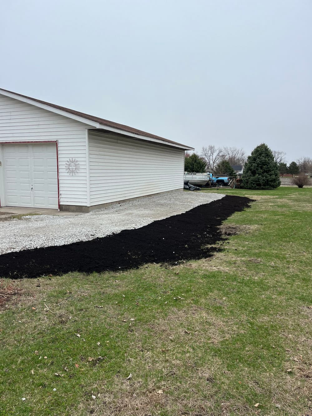 Freshly laid black mulch along a gravel driveway beside a white garage in a grassy yard.