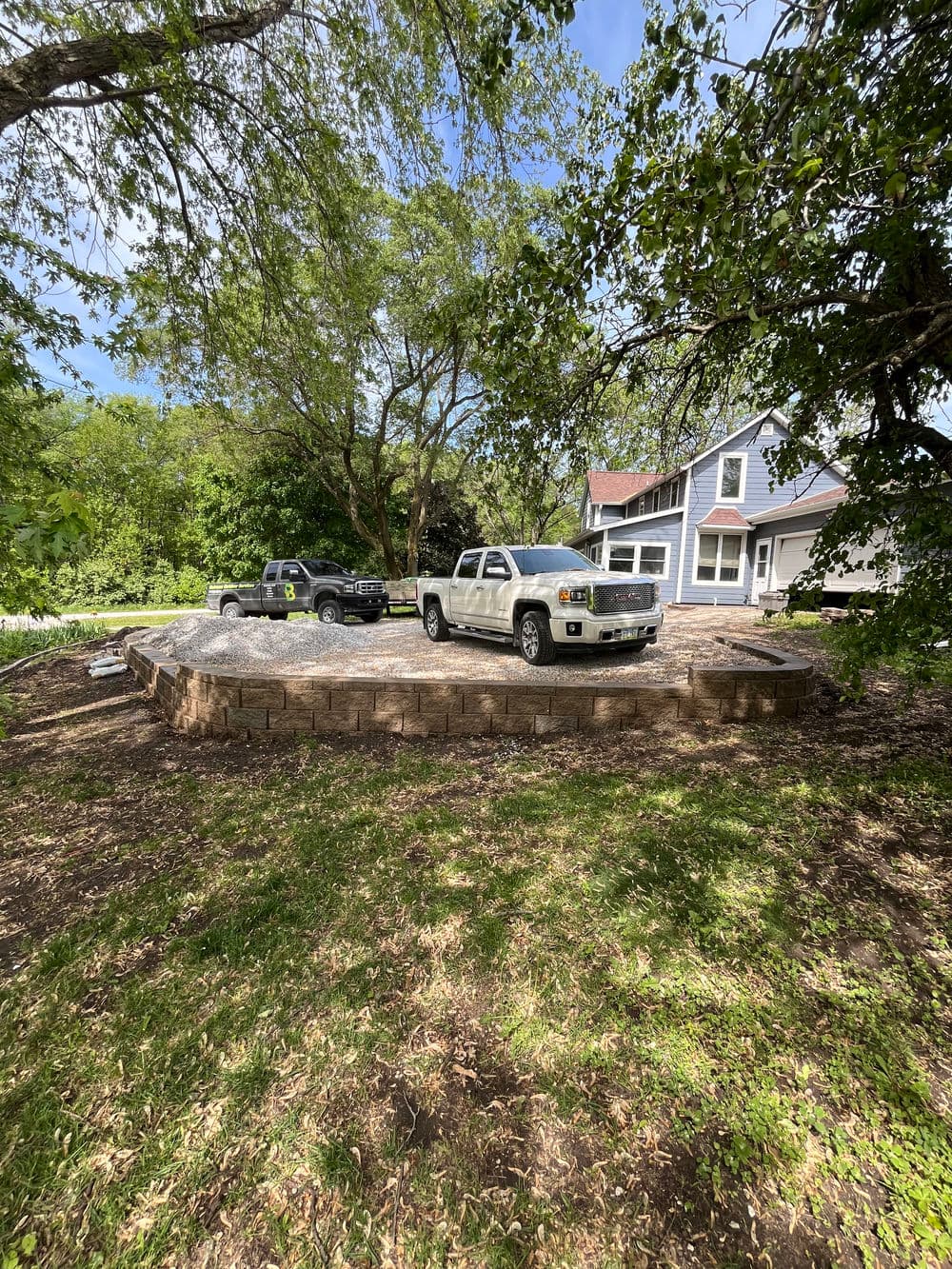 Two trucks parked on a new gravel driveway beside a house, surrounded by greenery.