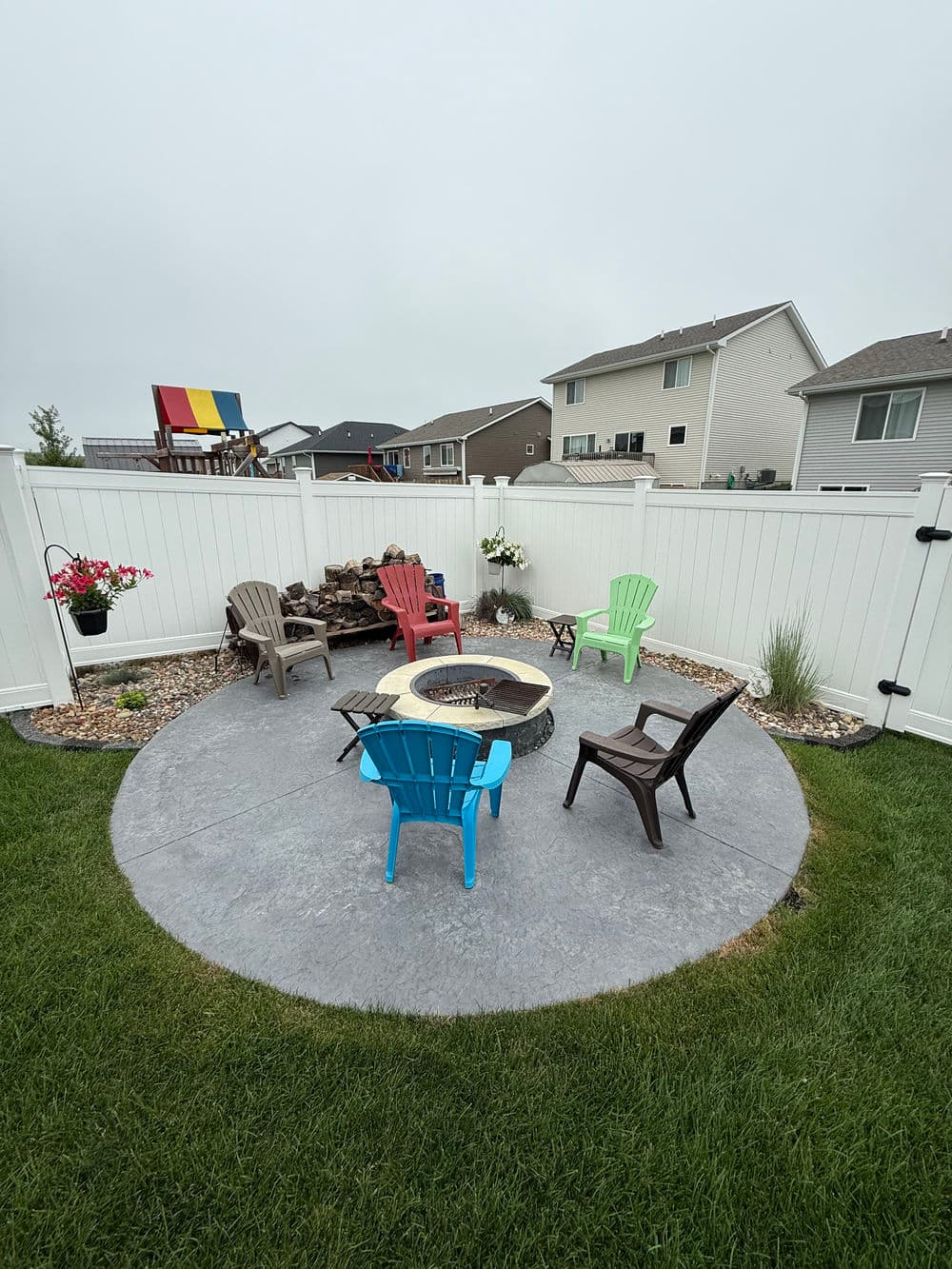 Backyard fire pit area with colorful chairs surrounded by a white fence and grassy lawn.
