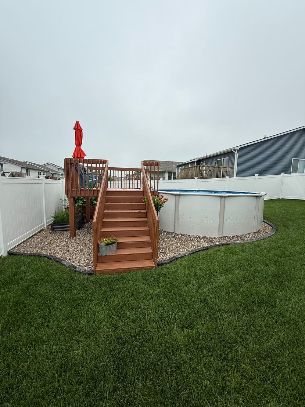 Above-ground pool with wooden deck and stairs, surrounded by grass and white vinyl fencing.