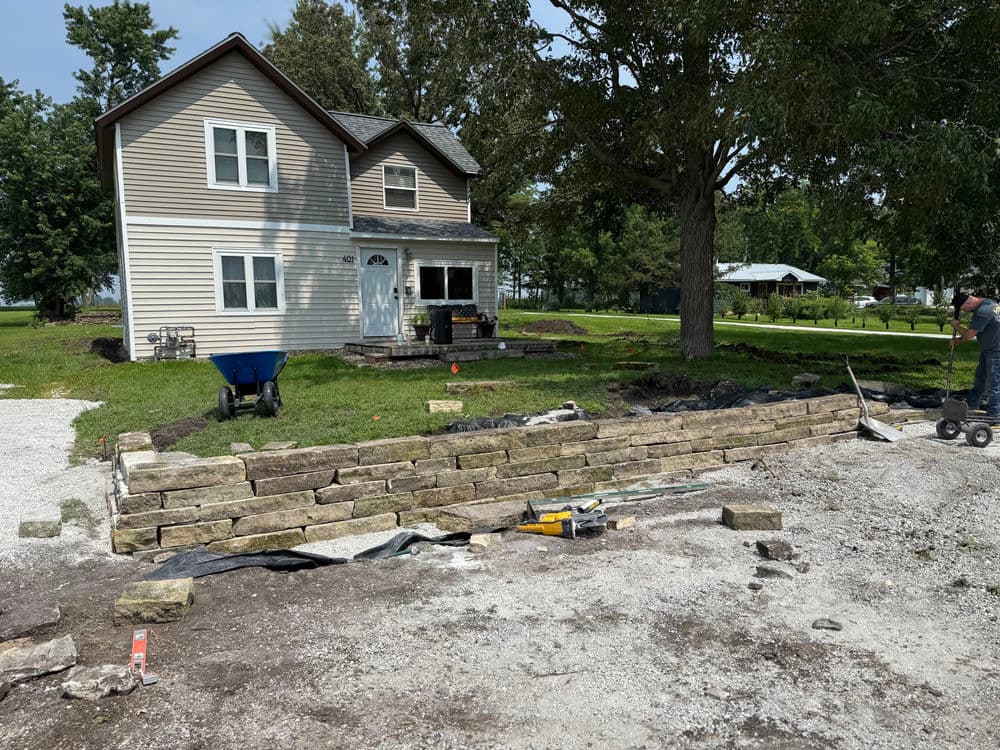 Stone retaining wall under construction in front of a house with landscaped yard and tools.